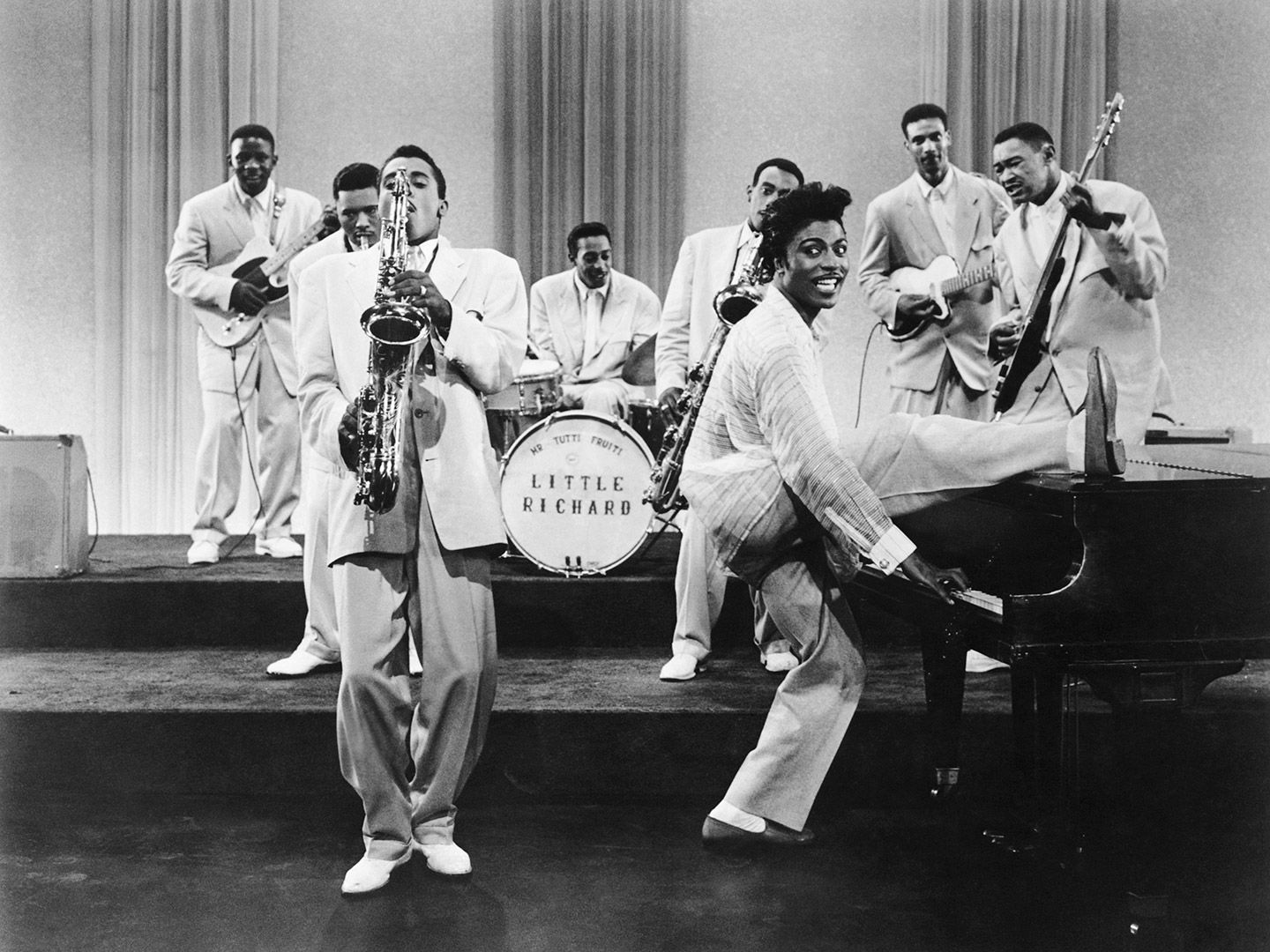 Little Richard plays the piano with one leg resting on the top of the piano.